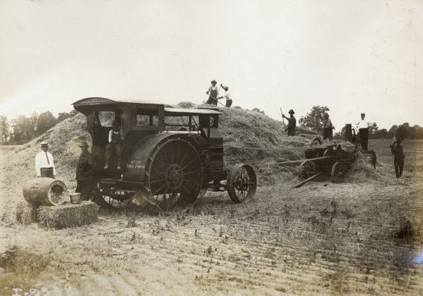 Workers on and near a large pile of hay use an International Harvester Mogul(?) 15-30 tractor and hay press in a field.