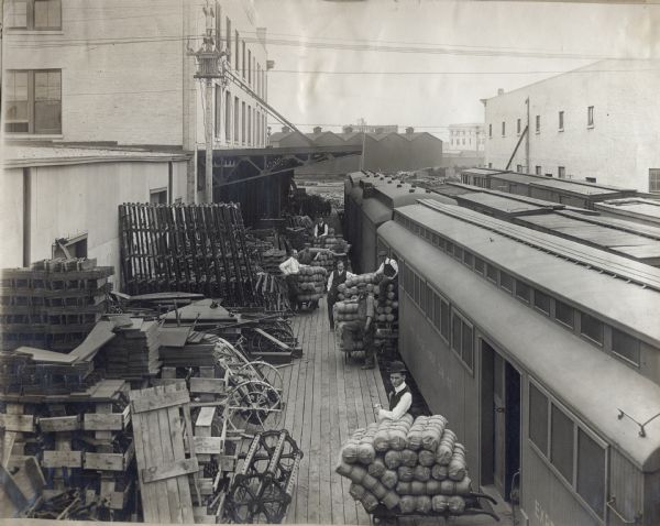 A group of men stand near rows of trains where crates, packages and pieces of machinery have been unloaded onto the platform. The platform is probably near an International Harvester branch house, possibly at Regina, Saskatchewan, Canada.