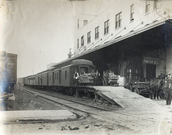 A group of men stand near a train at the loading dock of an International Harvester branch house, most likely at Regina, Saskatchewan, Canada. A horse-drawn wagon is parked near the dock.