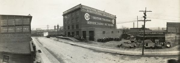 International Harvester Branch House. Signs on the building advertise agricultural implements, motor trucks and Kerosene engines and tractors. Many small tractors line the side of the building and a train docking station can be seen towards the back of the building.