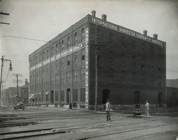 International Harvester branch house. A bystander watches a man work on one of the railroad tracks outside the building. An man in a automobile is driving up the street towards the railroad tracks.