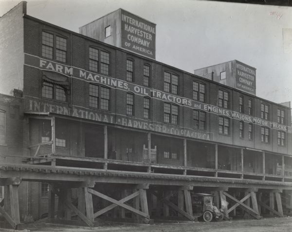 Exterior view of International Harvester branch house. Signs on the building advertise farm machines, oil tractors, engines, wagons and motor trucks. A long loading dock is above an elevated railroad track, and an automobile is parked underneath the tracks at ground level.