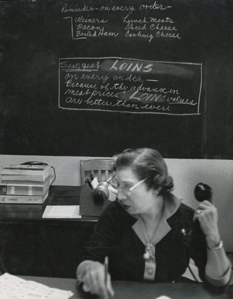 Gertrude Martin, operator for Hazan's Telephone Supermarket, at her desk with a phone in her hand. Behind her a chalkboard displays a reminder to "suggest loins on every order."