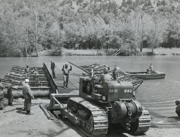 Soldier using an International TD-18 crawler tractor (TracTracTor) during a training exercise. The original caption reads: "A graduate of the school assigned to the Fifth Engineer Battalion shows he learned his lesson well. After pushing a stranded pontoon bridge section off pilings, he moves the TD-18 on board, floats to new site of bridge, helps install it."