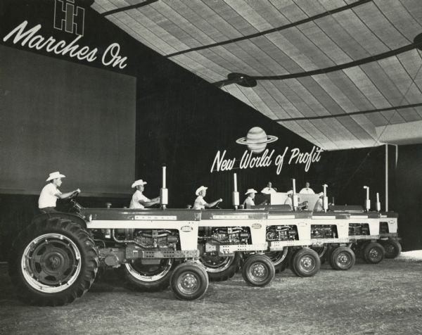 Men in cowboy hats show Farmall 460 tractors at an International Harvester product introduction show. Signs in the background read: "New World of Profit" and "IH Marches On."