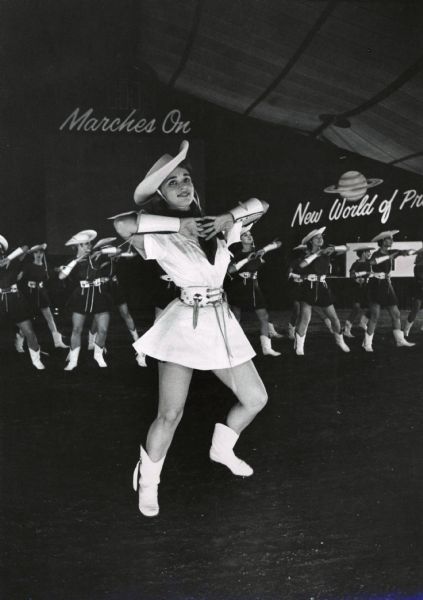 Kilgore Texas Rangerettes dancing at an International Harvester product introduction show. Sign's in the background read: "New World of Profit" and "IH Marches On".