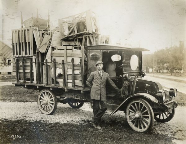 A man is standing in front of an International Model F (or 31) truck loaded with a variety of household items, including a metal washtub and wooden sled.