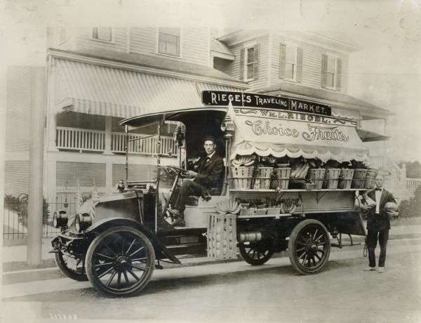 Two men stand near Riegel's Traveling Market truck, an International Model F (or 31) modified to hold baskets of bananas, pineapples, corn, lettuce, asparagus and many other kinds of fresh fruits and vegetables.