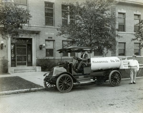 Two men pose with their International Model F (or 31) oil truck. The truck was operated by the Marshall Oil Company truck. A can on the back of the truck advertises for French Auto Oils, and the truck is parked in front of the Marshall Oil Company building.