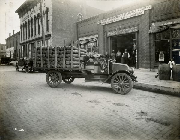 A man sits in a parked International Model F (or 31) truck filled with cylindrical packages outside of Studebaker Wagons and Buggies dealership, where a group of men stand at the entrance. The store also advertises for: the First Implement and Automobile Company, Automobiles and Farm Machinery, Fairbanks-Morse Gasoline Engines, Deering Farm Machines, Congress Tires, etc. Watsons Opera House is on the left.