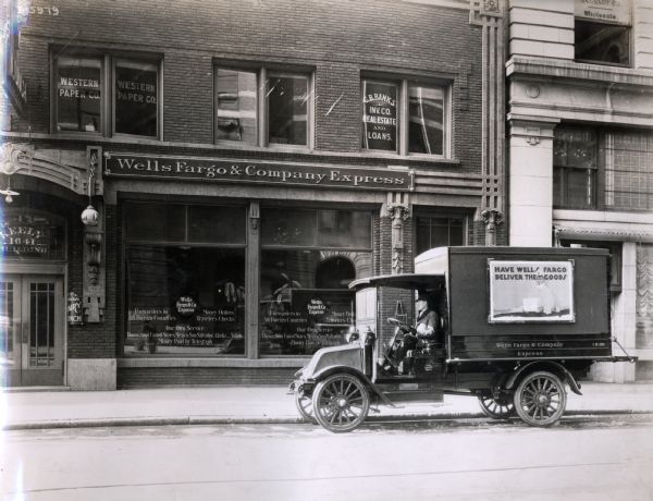 A man sits in an International Model F (or 31) truck operated by Wells Fargo and Company Express delivery services. The truck is parked outside the Wells Fargo and Company Express store in the Keely Building.