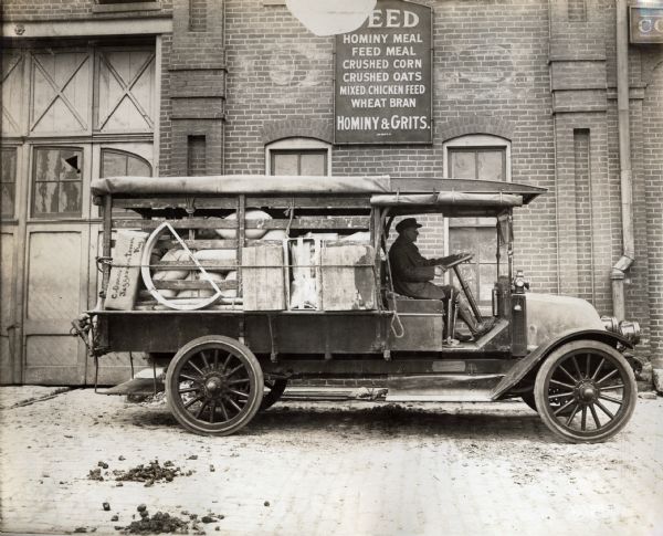 A man drives an International Model F (or 31) truck owned by a feed store. The truck is parked next to a building with a sign that reads: "Feed, hominy meal, feed meal, crushed corn, crushed oats, mixed chicken feed, wheat bran, hominy & grits."