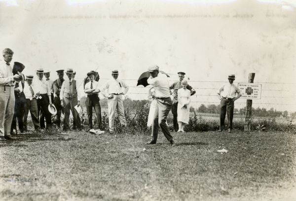 A small crowd gathers around a man as he tees off at a golf course.