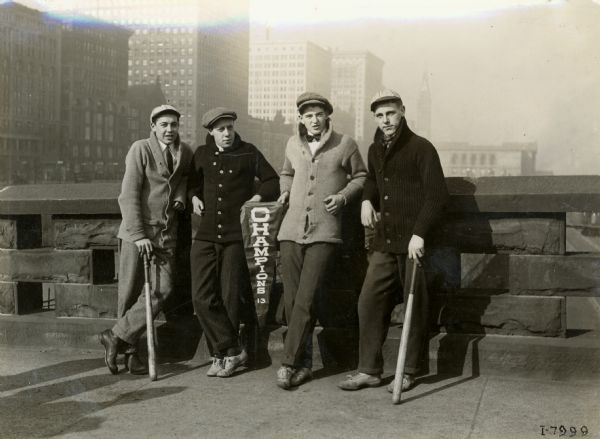 Four young men are stand up against a low stone wall displaying a small pennant with the word "Champions" on it. Two of the young men are holding baseball bats, and they may have been members of an International Harvester company baseball team. They appear to be standing on a city bridge, possibly in Chicago.