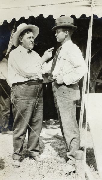 Two men standing near a display tent at a tractor exhibition. One man appears to be talking animatedly while the other listens.