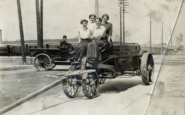 Four women are sitting on an International Harvester manure spreader. The women are identified as "Madison Girls," most likely employees at International Harvester's Madison, Wisconsin offices. The photo appears to have been taken in front of the company's branch house building at 301 S. Blount Street. Behind them on the left is a man driving a truck.