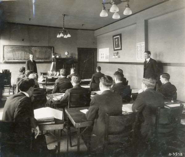 A group of men sit at desks and listen to J. Newman from Deering Works give a lecture on electricity.