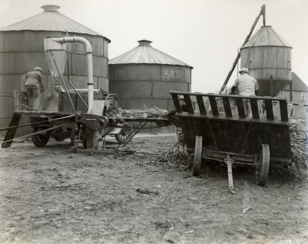Two men use a No. 3 McCormick and Deering Hammer Mill in a farm yard. One man is standing in a wagon and the other is on the bed of a truck.