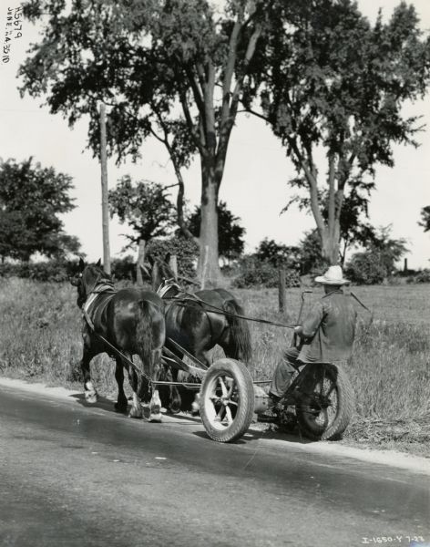 A man using a horse-drawn No. 7 mower with rubber tires to mow grass along a road in Hamilton, Ontario.