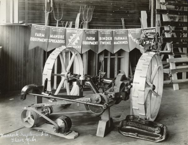 Mower and assorted mower parts on display in front of banners advertising McCormick-Deering farm equipment, twine, machines and motor trucks in Hancock Implement Co. Hancock Implement was an International Harvester dealership.