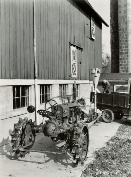 Men working with a Farmall F-30 tractor, a No. 1-B hammer mill and a wagon near a barn. The F-30 tractor has electric lights.