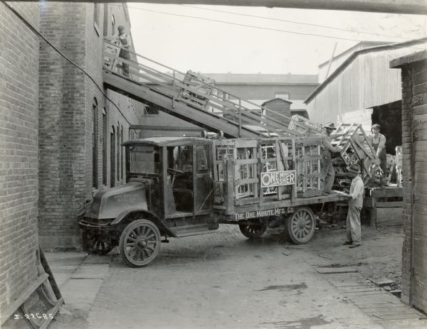 An International Harvester Model G-61 truck owned by the One Minute Manufacturing Co. carrying a shipment of One Minute Washers. The truck is parked in an urban alley at a second-story loading dock while men handle crates.