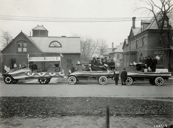 Three Model G-61 trucks are decorated for an International Harvester parade. A group of men, women and children sit in the middle car waving American flags, while a group of men simply stand in the third car.