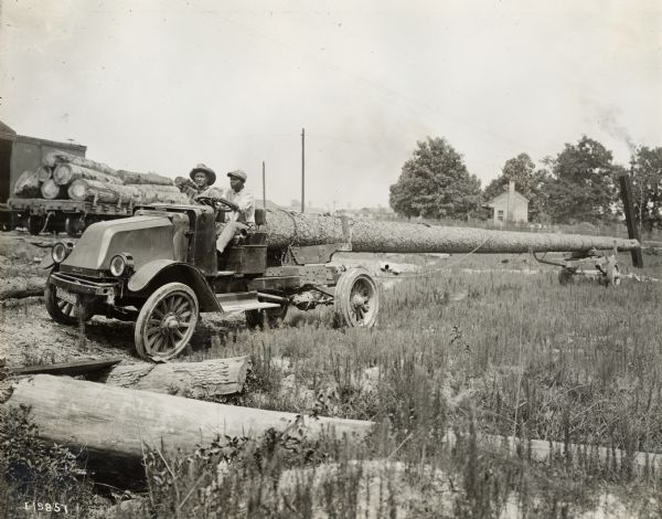 Two men hauling large logs with an International G-61 truck. Railroad flat cars loaded with logs are in the background.
