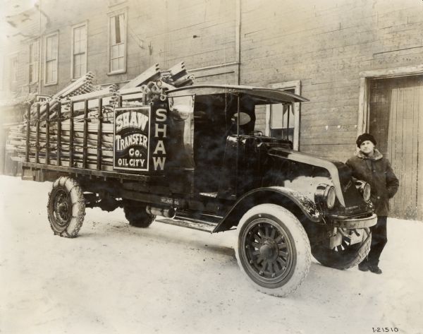 A driver is leaning against an International G-61 truck operated by the Shaw Transfer Company. The truck is parked on a snow-covered street and appears to be loaded with wooden ladder sections.