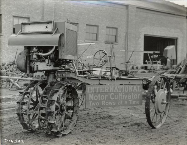 An experimental motor cultivator parked outside an International Harvester factory. A sign hanging from the cultivator reads: "International Motor Cultivator Two Rows at a Time".