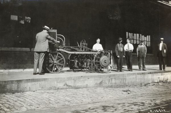 A man in a suit looks over an experimental motor cultivator while other men look on.