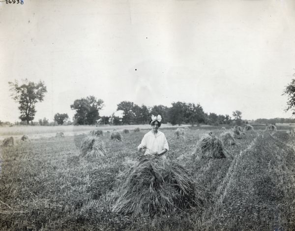 A woman with a large white bow in her hair stands behind shocks of grain in a field. She holds pieces of grain in her mouth and hands.