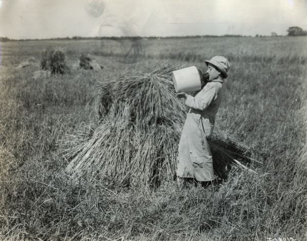 A small boy in a cap drinks from a heavy water jug near shocks of grain in a field.