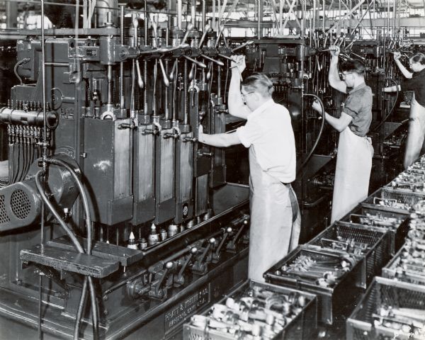 Factory workers at their machines in International Harvester's Chatham Works. Boxes of metal parts are lined up behind them.