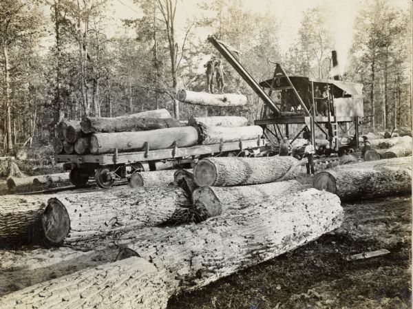 Two men are standing on a log suspended from a steam powered crane at an International Harvester logging operation. Large logs are in the foreground.