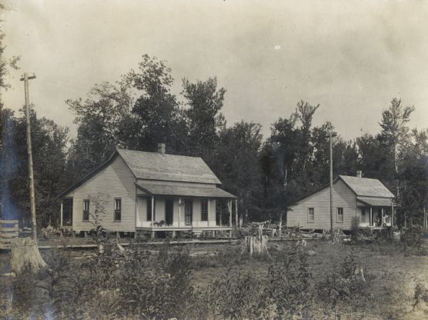 Two buildings, including the office of Dr. A.B. Allston (left), at an International Harvester logging camp or sawmill. People can be seen standing on the porch of the building on the right.
