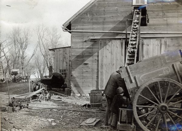 A father and young son loading corn from a wagon to a barn. A stationary engine is in the background.