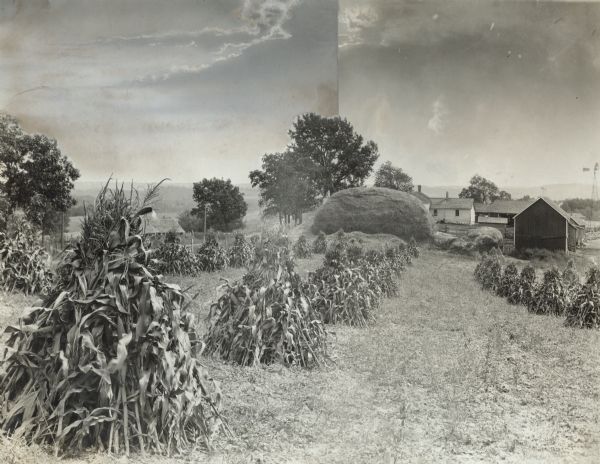 Farm with corn shocks and a large pile of hay. In the background are farm buildings and a windmill.