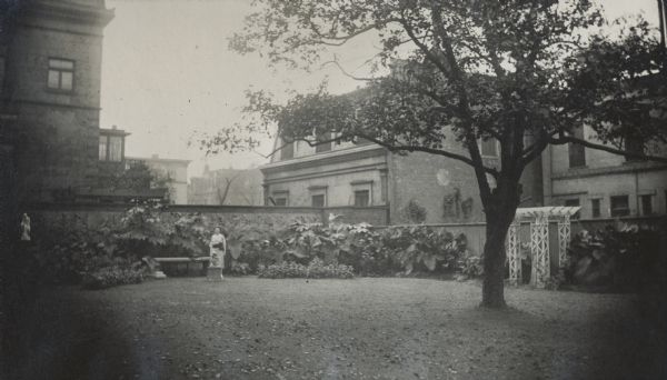 A woman is standing in the garden by the corner of 675 Rush Street, the residence of the McCormick family, which takes up an entire block between Erie and Huron Streets. The original caption indicates that this photograph was sent with a letter from William V. O'Brien to Nettie Fowler McCormick on September 7, 1921. Another note on the photograph reads: "Corner of 675 Rush St., garage, Huron St. house seen over garden on Michigan Avenue, between Huron and Erie Streets."