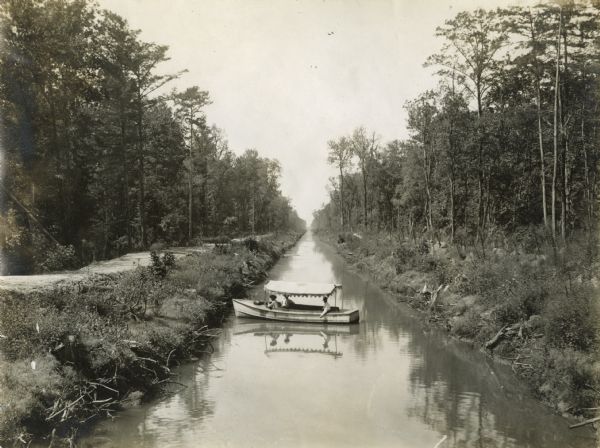 Three men in a covered boat on an International Harvester logging(?) canal surrounded by trees on either side.