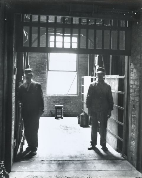 Two factory workers standing on a freight elevator, silhouetted by light from an open window behind them. The men probably worked at International Harvester's McCormick Works.