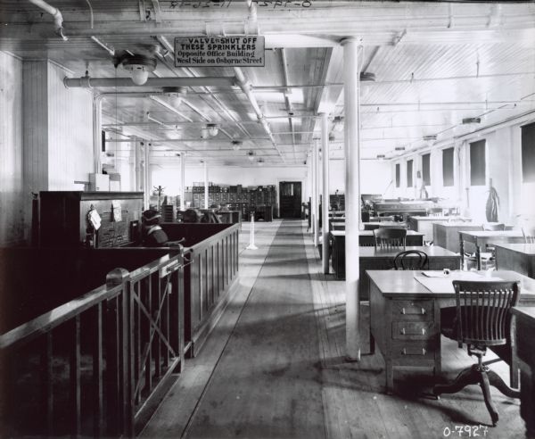 Offices at International Harvester's Osborne Works (later known as "Auburn Works"). The offices are empty except for a lone switchboard operator. A sign hanging from the ceiling reads: "Valve to shut off these sprinklers opposite office building west side on Osborne Street."