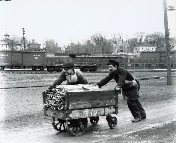 Two men demonstrate the "old way" of hauling using a push cart loaded with 2,200 pounds. The men are likely factory workers at Osborne Works (also known as "Auburn Works").
