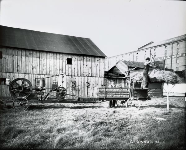 Two men demonstrate a motorized hay press in front of a barn and a Champion Works factory building.