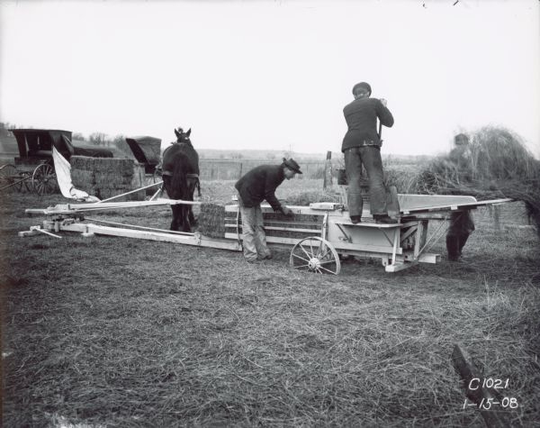 Three men in a field use a horse-powered hay press. Two carriages are on the the left.