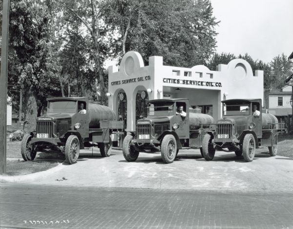 Three men in International Harvester oil trucks are lined up in front of a Cities Service Oil Co. building.