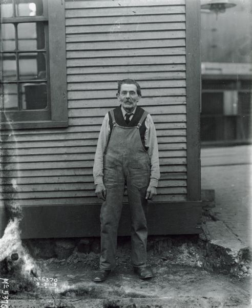 Full-length portrait of a man standing outdoors in front of a building. He is wearing eyeglasses, and overalls over a shirt, vest and necktie.