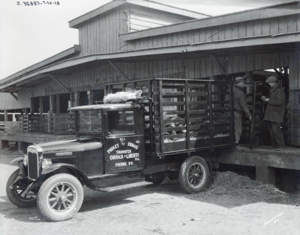 International truck with "Padget and Frankes Transfer, Orrick or Liberty MO." painted on the door. Two pigs are in the trailor, which is parked at a loading dock. Three men are standing on the loading dock.
