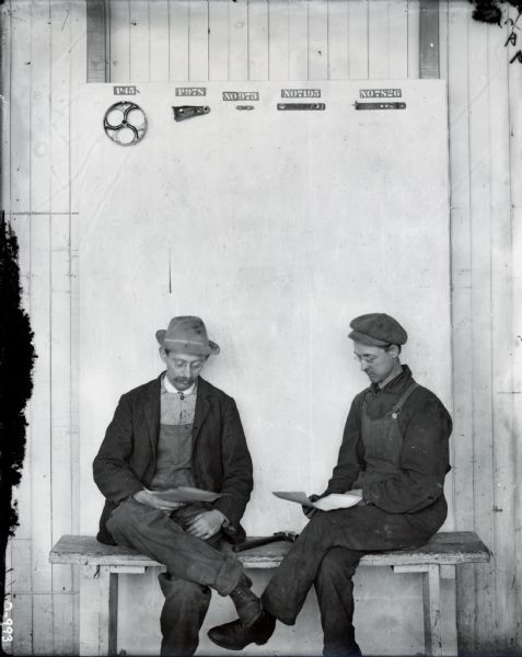 Two men pause to read parts lists(?) while mounting farm equipment parts to a large board. The parts are labeled with part numbers. The board was probably used to produce parts lists and diagrams for catalogs and operator's manuals. The men are most likely employees at International Harvester's Osborne Works (the factory was later known as Auburn Works).