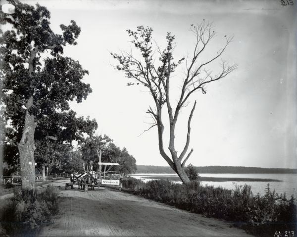 View down road towards a man and a dog standing next to a horse-drawn McCormick grain binder near a lake.
